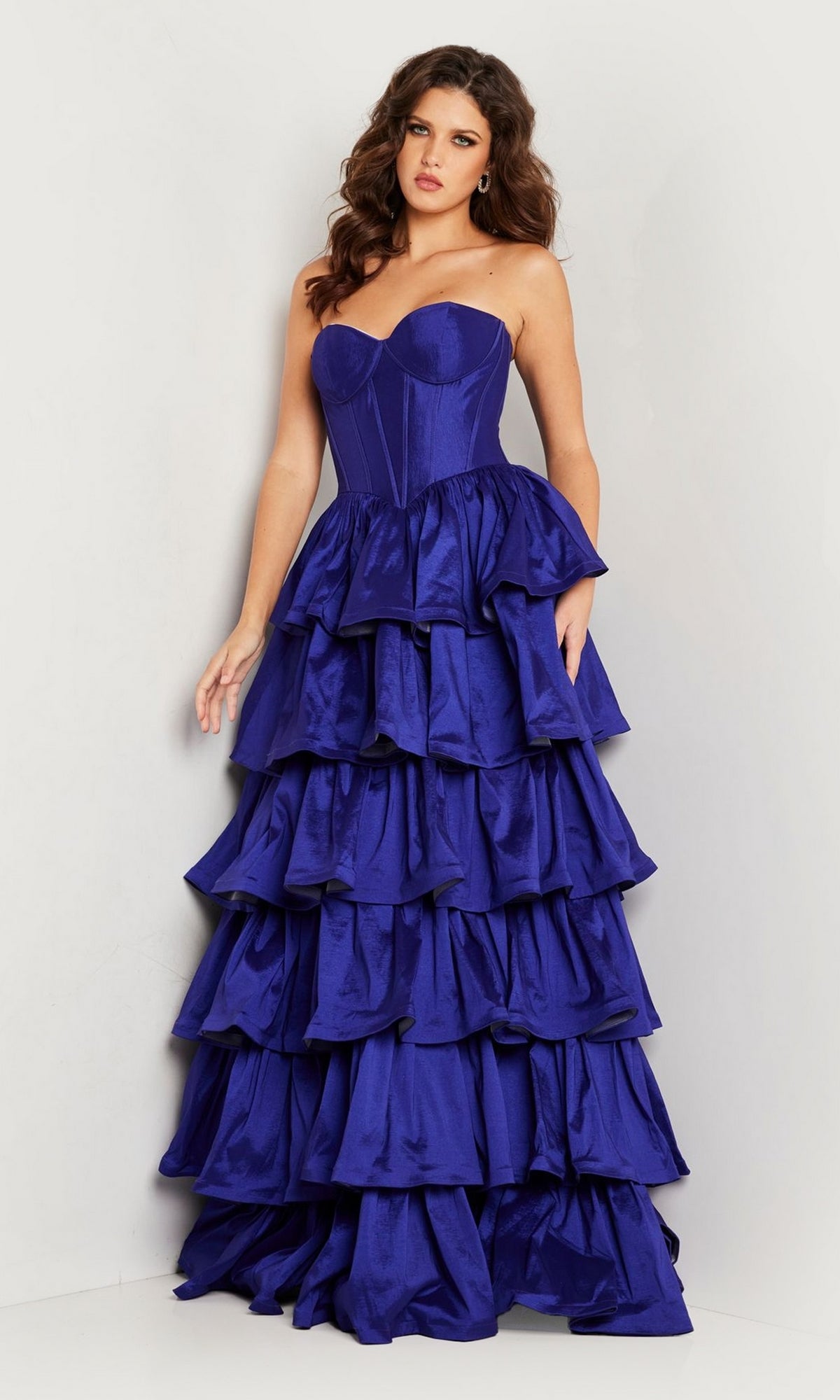 Against a plain background, a woman wears the Jovani 36619—a strapless, royal blue prom ball gown featuring elegant tiered ruffles and a fitted bodice.
